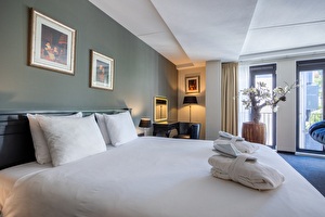 Junior Suite in Amrâth Grand Hotel Frans Hals met twee eenpersoonsbedden, een zwarte stoffen fauteuil, rode bewerkte gordijnen, een spiegel in de hoek van de kamer en kunst boven het bed