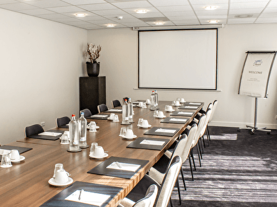 Meeting room with a long wooden table set up with notepads, pens, cups, and water bottles, featuring a projection screen on the wall. A flip chart with a welcome message is placed in the corner.