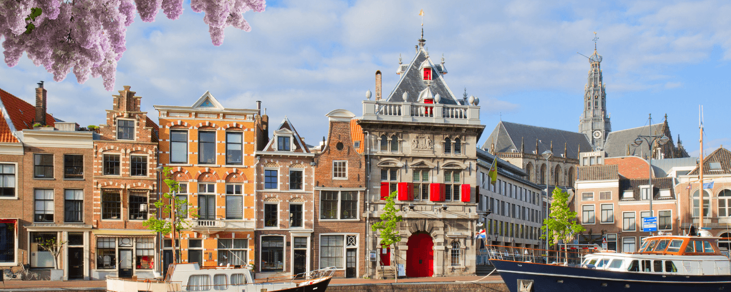 Schilderachtige gracht in Haarlem met historische gebouwen, een gotische kerk en aangemeerde boten. Lila bloesems hangen in de buurt onder een deels bewolkte lucht.