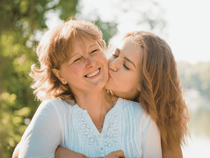Laughing mother lovingly embraced and kissed on the cheek by her daughter during a sunny day in a green, natural environment.