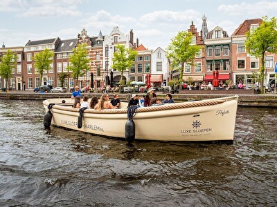 A luxury boat filled with passengers cruising through the canals of Haarlem, with historic buildings and outdoor terraces in the background.