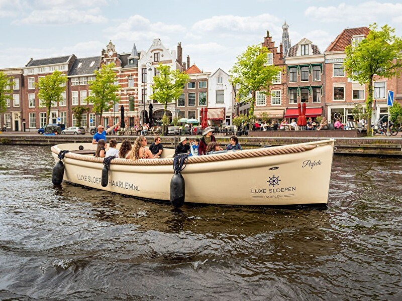 A boat with people sails through a picturesque canal, surrounded by green trees and historic buildings.