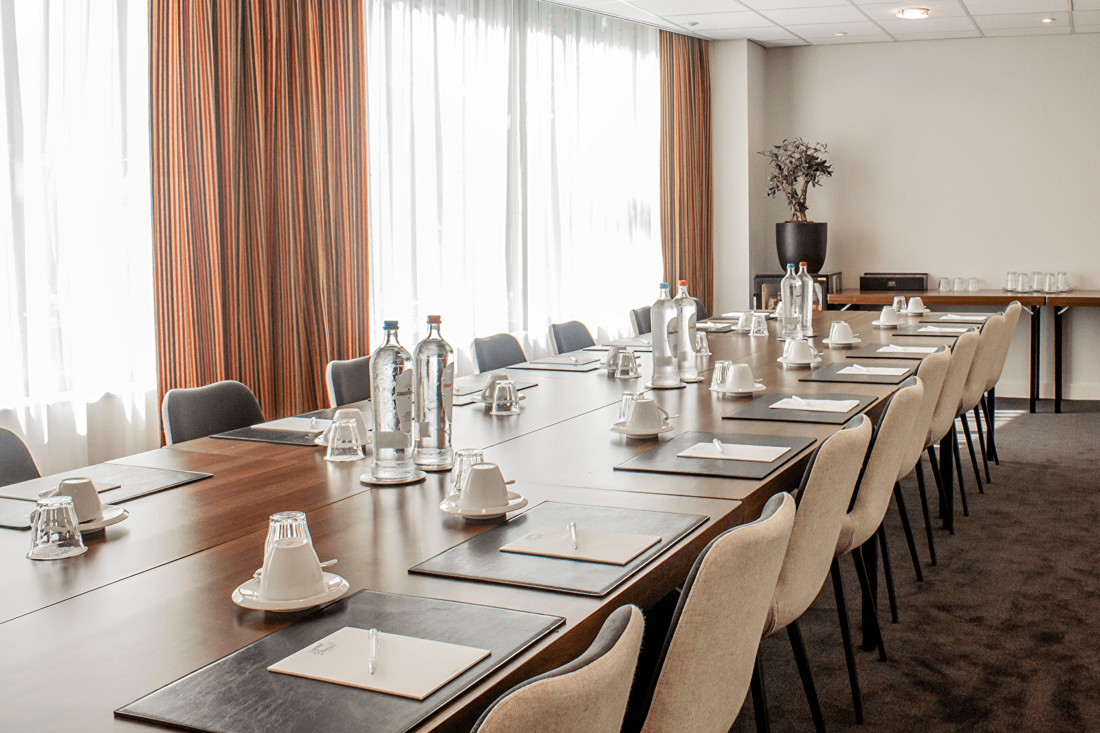 Vergaderzaal in Amrâth Grand Hotel Frans Hals met lange opstelling, comfortabele stoelen, daglicht via grote ramen en vergaderfaciliteiten zoals notitieblokken, pennen en waterflessen.